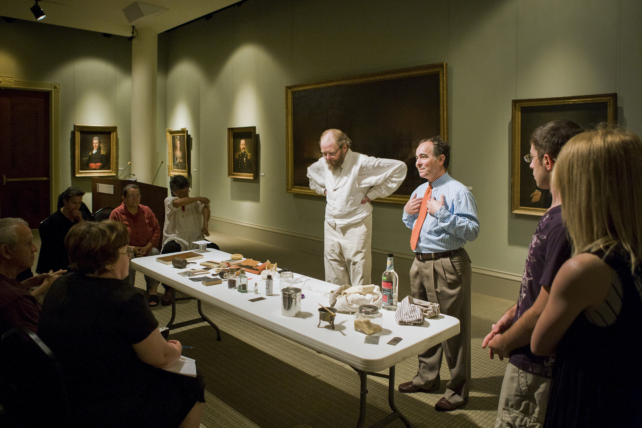 Two men give a presentation while standing behind a table full of historical objects, talking to a crowd.