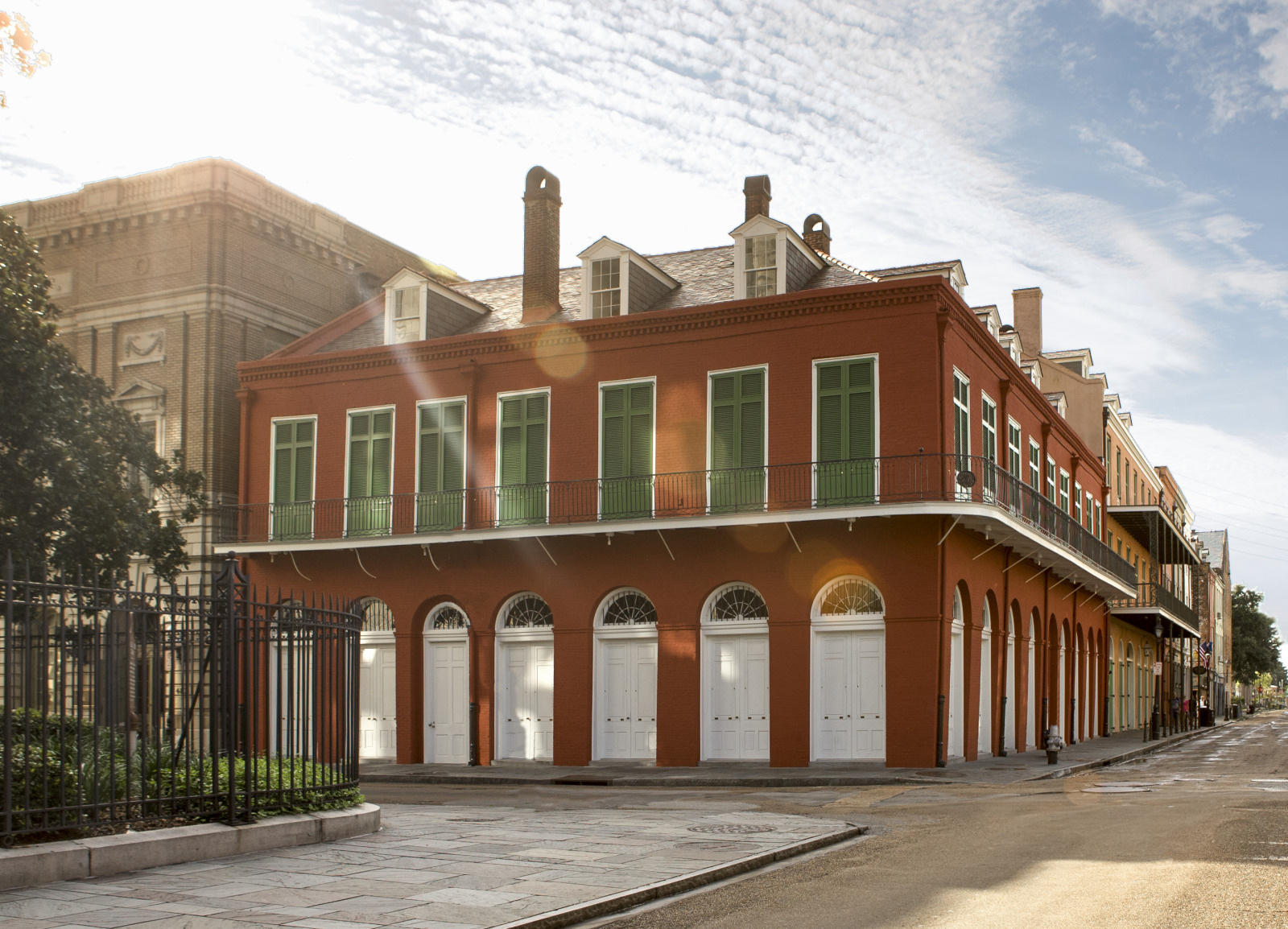 A two-story red building with green shutters and white doors stands on a street corner under a bright blue sky with scattered clouds. A wrought-iron fence and trees are visible on the left, with sunlight casting a lens flare across the scene.