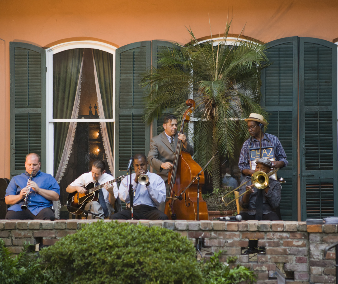 A jazz band performs on a patio in front of green shutters and an orange wall. The group includes a trumpet player, guitarist, double bassist, and two other musicians, creating a lively outdoor ambiance. Plants and shrubbery are in the foreground.