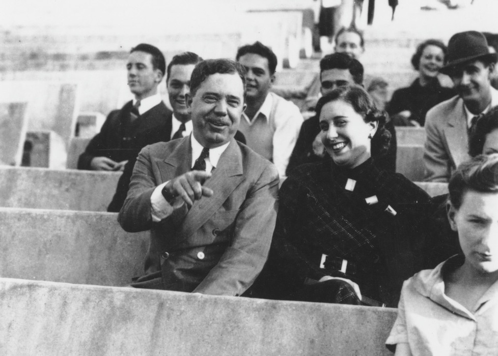A black and white photo of people sitting in outdoor stadium seats. A man in a suit is pointing and smiling, sitting next to a woman who is also smiling. Others around them are engaging and looking in various directions.