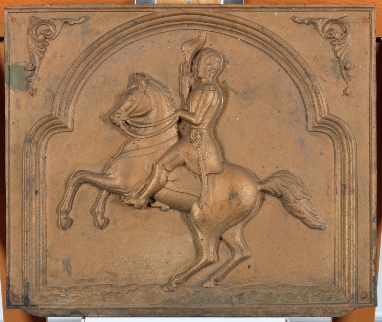 A relief sculpture depicting a figure on horseback holding a hat, set within an ornate rectangular frame. The horse is rearing up, and the scene is crafted in a metallic, weathered bronze tone.