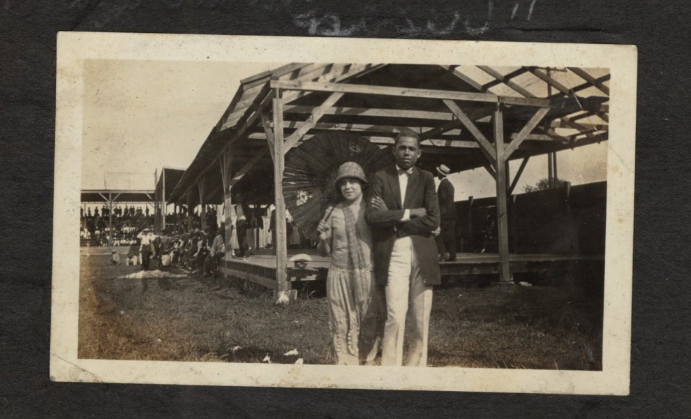 A vintage photograph of a woman and man standing in front of a wooden grandstand. The woman is wearing a dress and hat, holding a parasol, while the man is in a suit. A crowd is seated in the background.