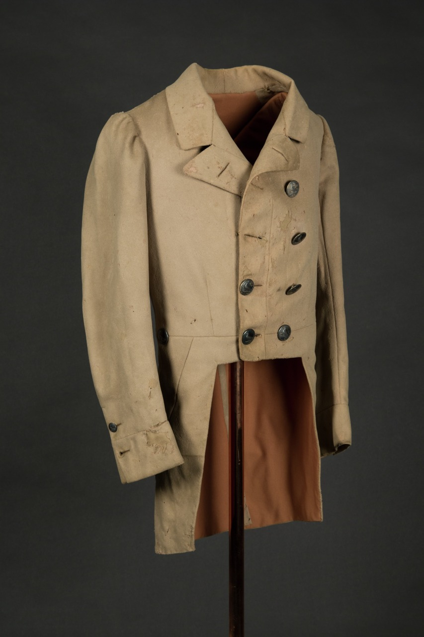 A vintage tan tailcoat with black buttons, displayed on a stand against a dark background. The coat features a high collar and long sleeves, with visible wear and distress, highlighting its historical nature.