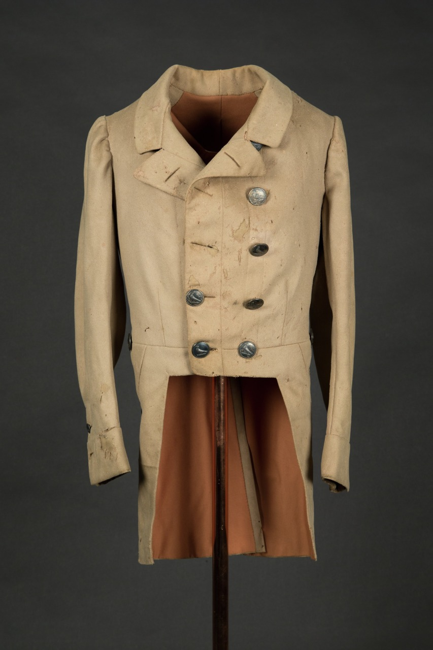 A vintage beige coat with brass buttons displayed on a stand against a dark background. The coat is long with tails, showing signs of age and wear.
