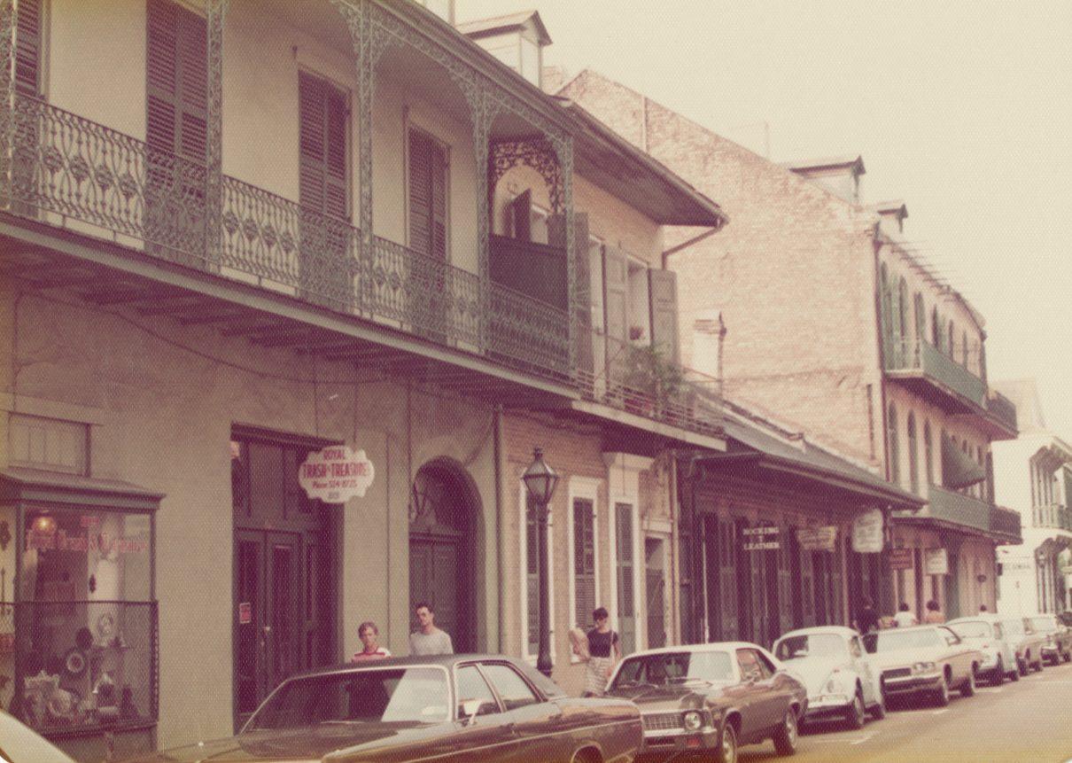 A vintage street scene with cars parked along a row of historic buildings. The architecture features ornate iron balconies. People are walking on the sidewalk, and a sign for French Quarter Restaurant is visible. The overall tone is nostalgic and historic.