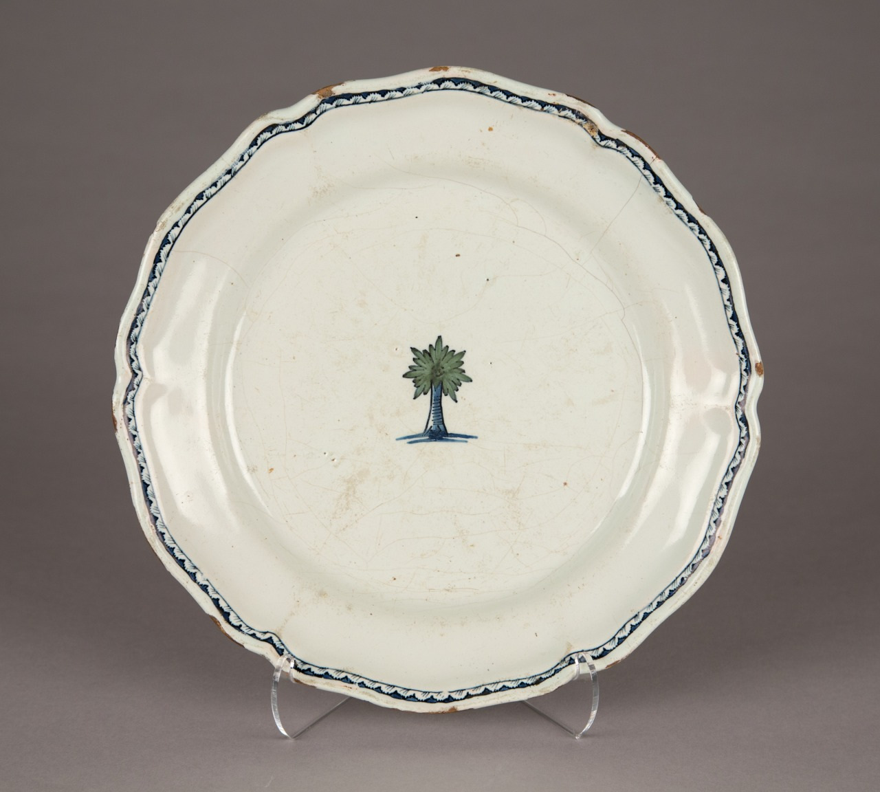 A round ceramic plate with a scalloped edge and a simple design. The center features a small green tree with a blue trunk. The plate has a delicate blue border near the rim and shows signs of wear.