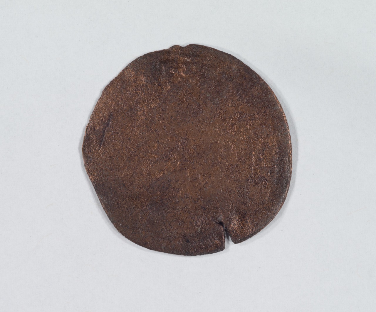 A round, weathered copper coin with a smooth, worn surface is displayed against a plain light background. The coin shows signs of age and has a small notch near its edge.