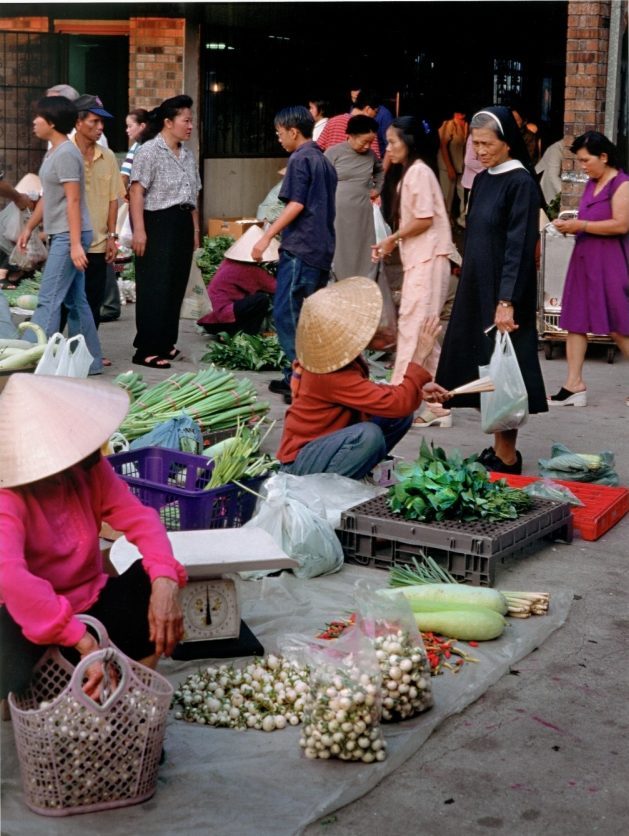 A bustling outdoor market scene: vendors sit on the ground selling vegetables and herbs, including leafy greens, peppers, and gourds. Shoppers walk by, some holding bags. A woman in traditional Vietnamese attire and a nun are visible.
