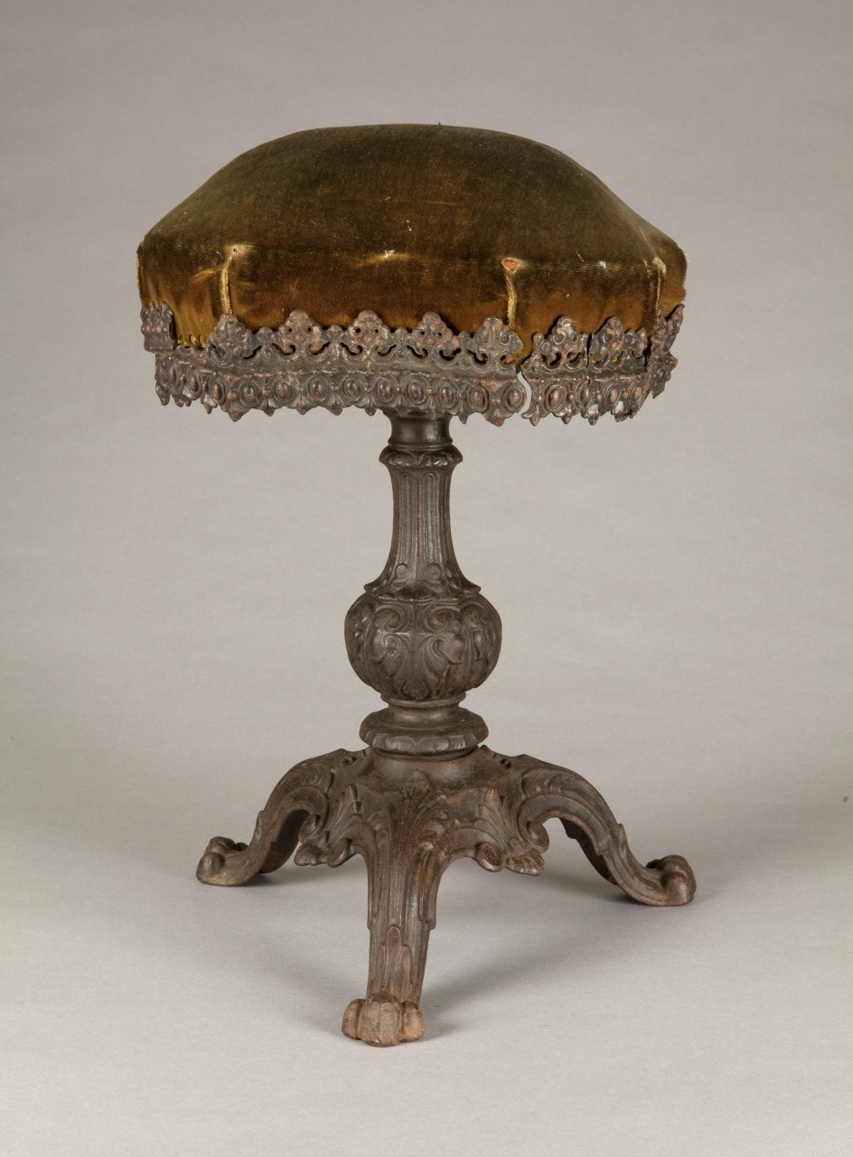 An ornate antique stool with a velvet seat and intricate carvings on the wooden base and legs, set against a plain backdrop.