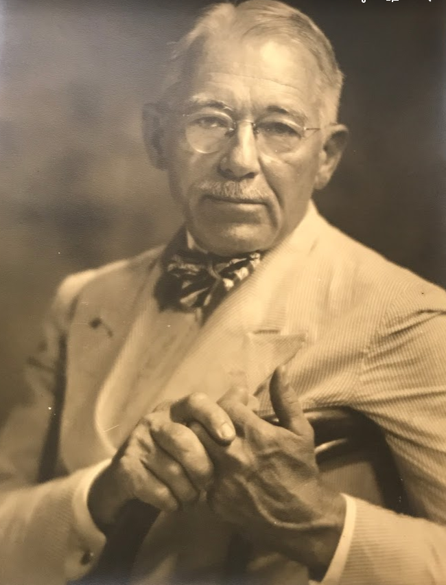 A distinguished older man with glasses and a mustache is wearing a suit and bow tie. He is holding a cane in both hands. The photo is in sepia tone, giving it a vintage look.