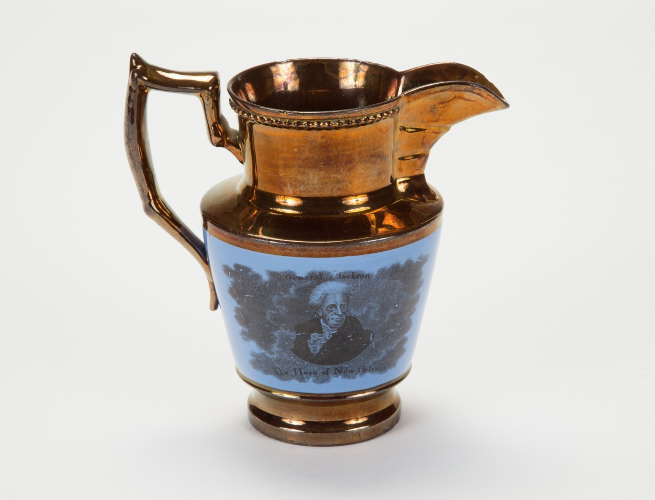 A vintage copper jug with an ornate handle and spout, featuring a blue and black portrait of a man on its side. The jug has a shiny, reflective surface and intricate detailing around the rim.