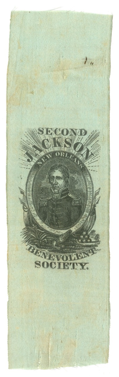 A vintage ribbon featuring a black-and-white illustration of a man in a military uniform, surrounded by stars and laurel leaves. The text reads Second Jackson Regiment Benevolent Society. The ribbon is light green with slight discoloration.