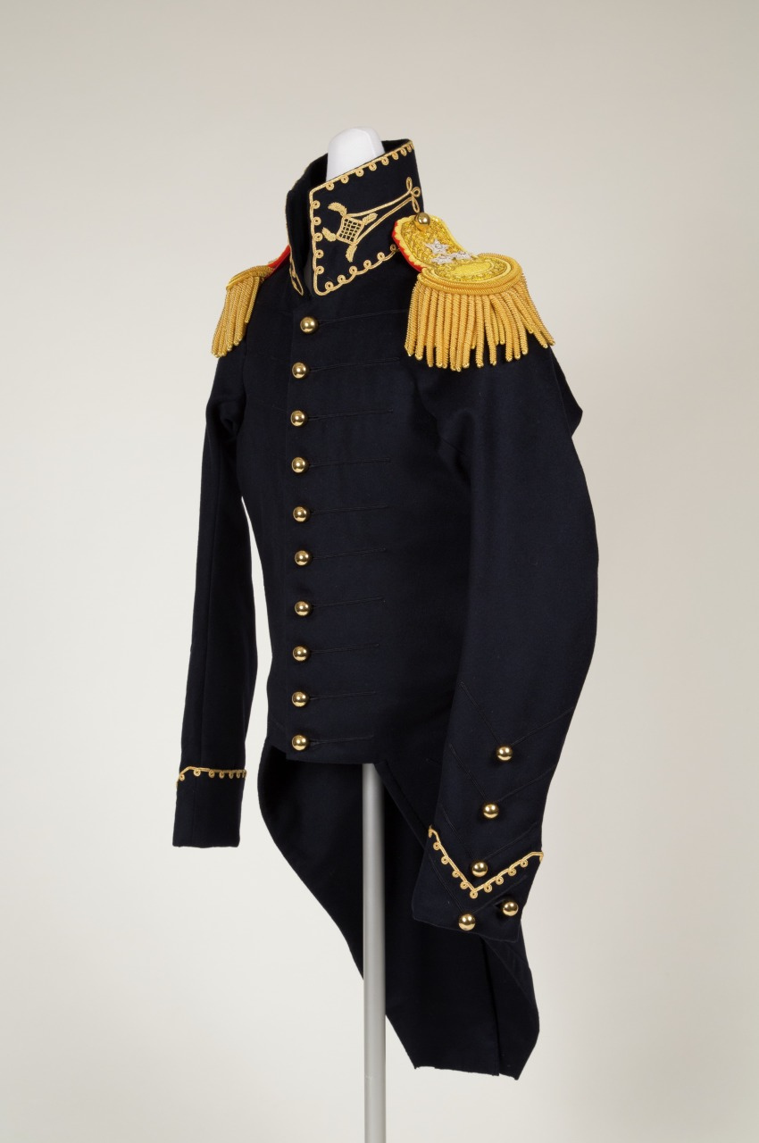 A historical military uniform coat with gold epaulettes and decorative gold buttons on a mannequin. The coat is dark navy with gold trim and intricate detailing on the epaulettes. The mannequin is positioned against a plain background.