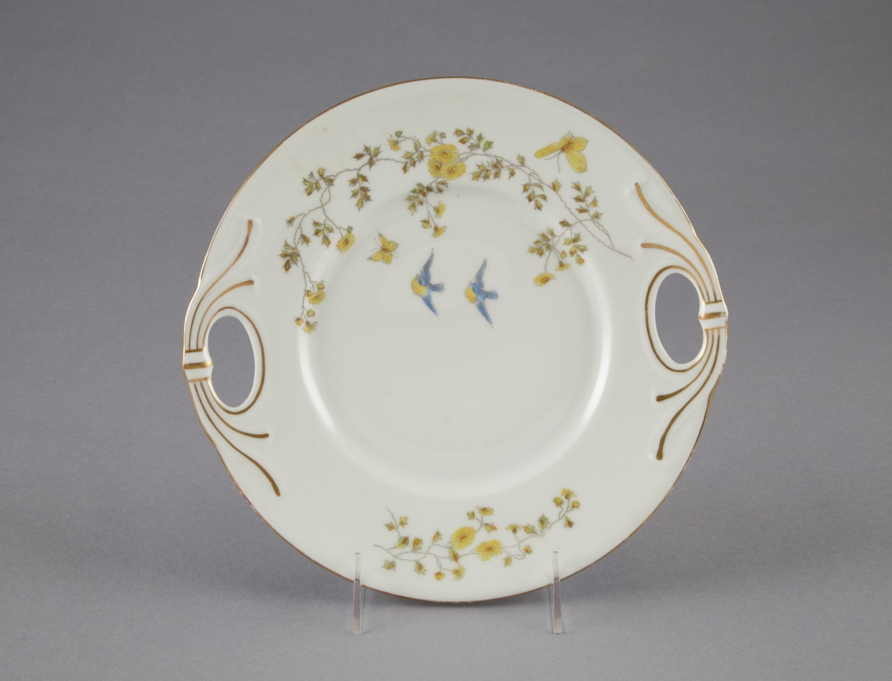 A round white plate with delicate yellow flowers and two blue birds painted on it. It has two decorative cut-out handles and gold rim accents, placed against a gray background.