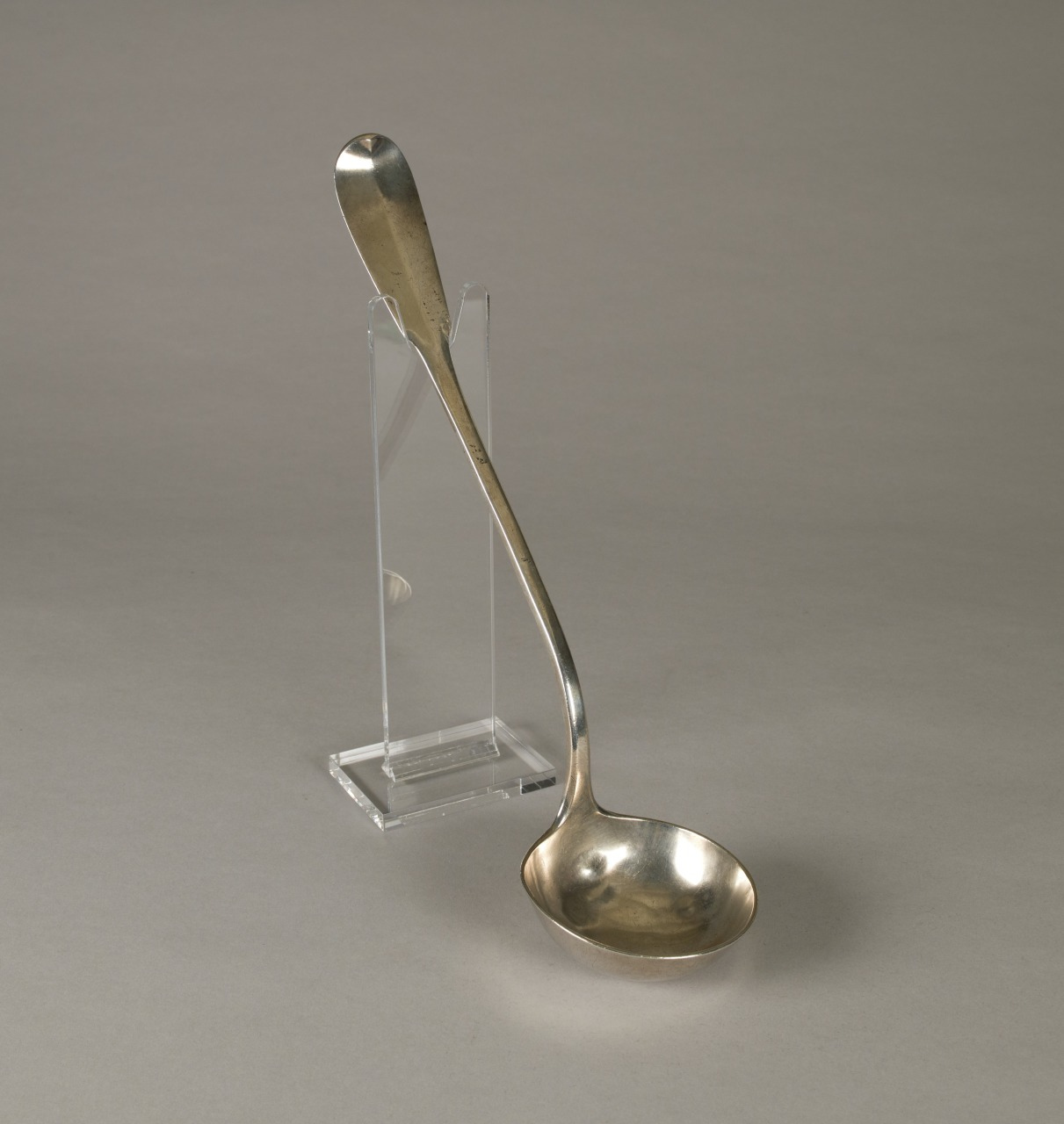 A silver ladle with a long handle and a deep, round bowl is displayed upright on a transparent stand against a plain gray background.