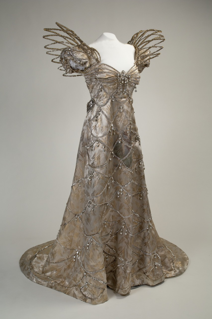 An elegant gown with ornate silver fabric, featuring structured, decorative elements on the shoulders and intricate beadwork forming a pattern across the bodice and flowing skirt.