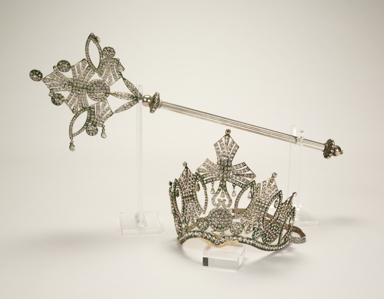 A detailed silver scepter with ornate designs is displayed alongside an intricate silver crown featuring jewel accents. Both items are set on a clear stand against a neutral background.