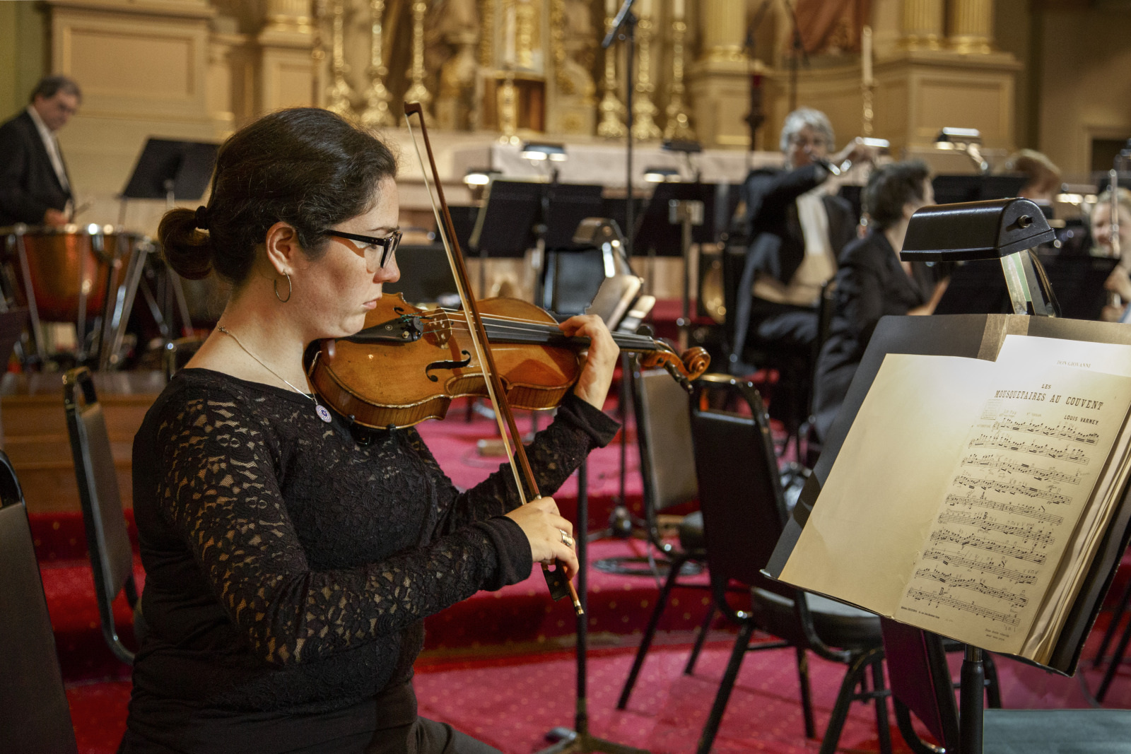 A woman wearing glasses and a black lace top plays the violin in an orchestra. She is seated with sheet music in front of her. Other musicians with various instruments are visible in the background, set in a formal performance hall.