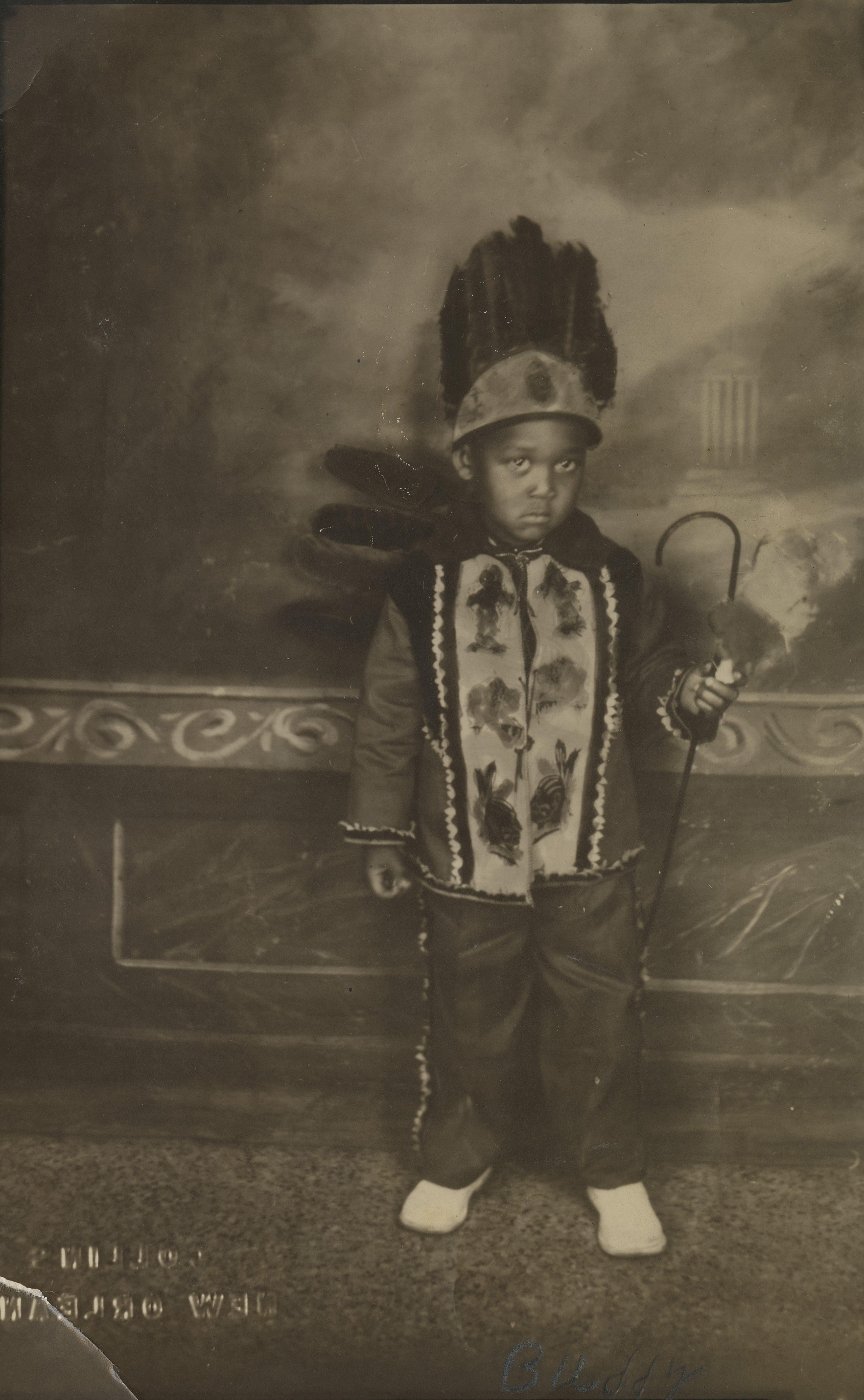 A young child in a festive costume stands against a painted backdrop. They wear a headdress with feathers, a decorated jacket, and hold a curved cane. The sepia-toned photo gives a vintage feel.