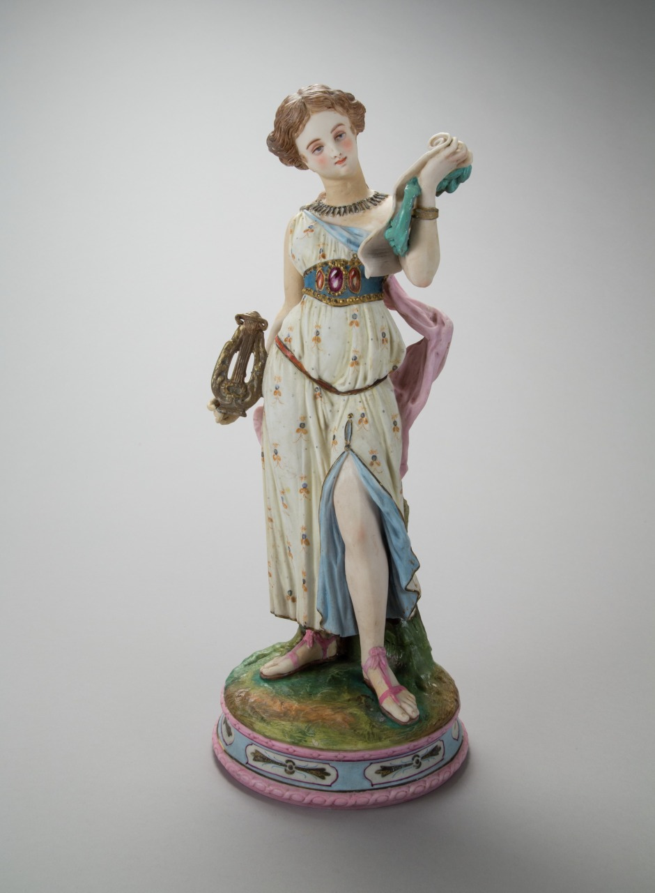 A porcelain figurine of a woman in a classical dress, standing elegantly. She holds a scroll in one hand and a lyre in the other. Her dress is adorned with floral patterns and has a blue sash. The figure stands on a decorated base.