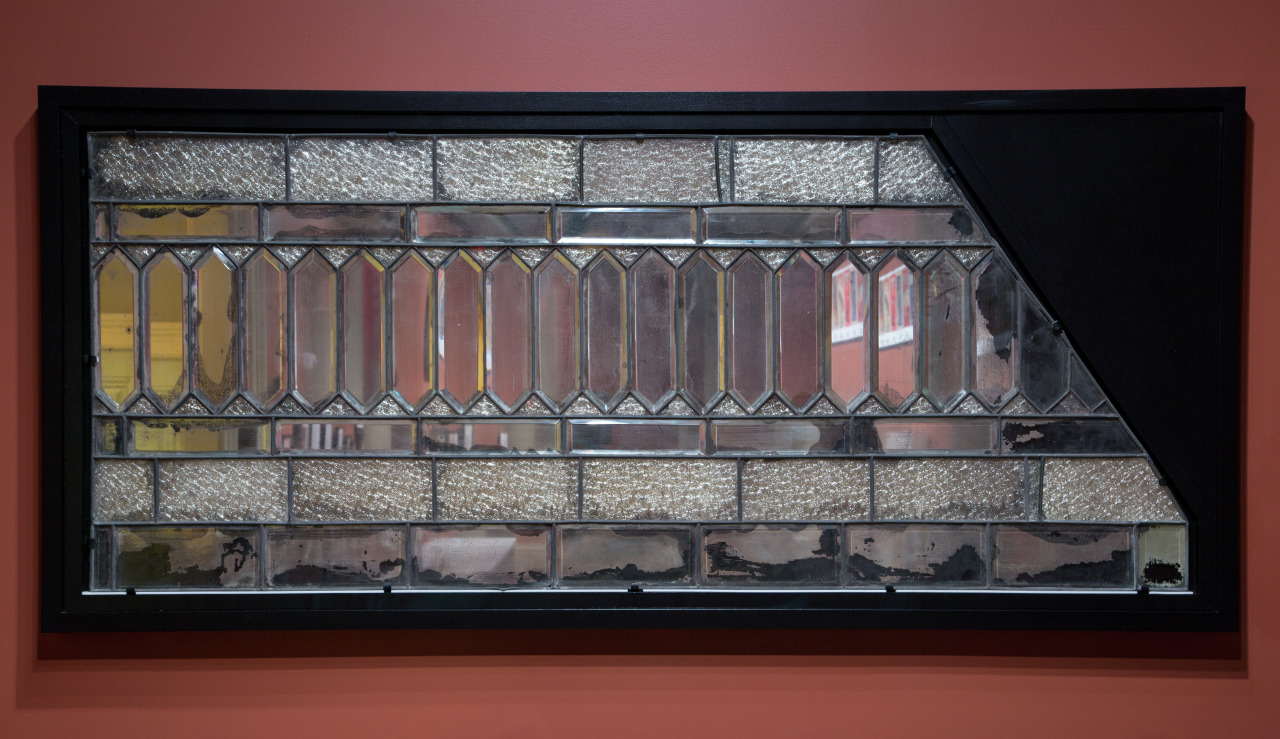 A rectangular stained glass artwork in a black frame against a red wall. The design features textured glass panels in a geometric pattern with horizontal and vertical lines, creating a reflective and intricate appearance.