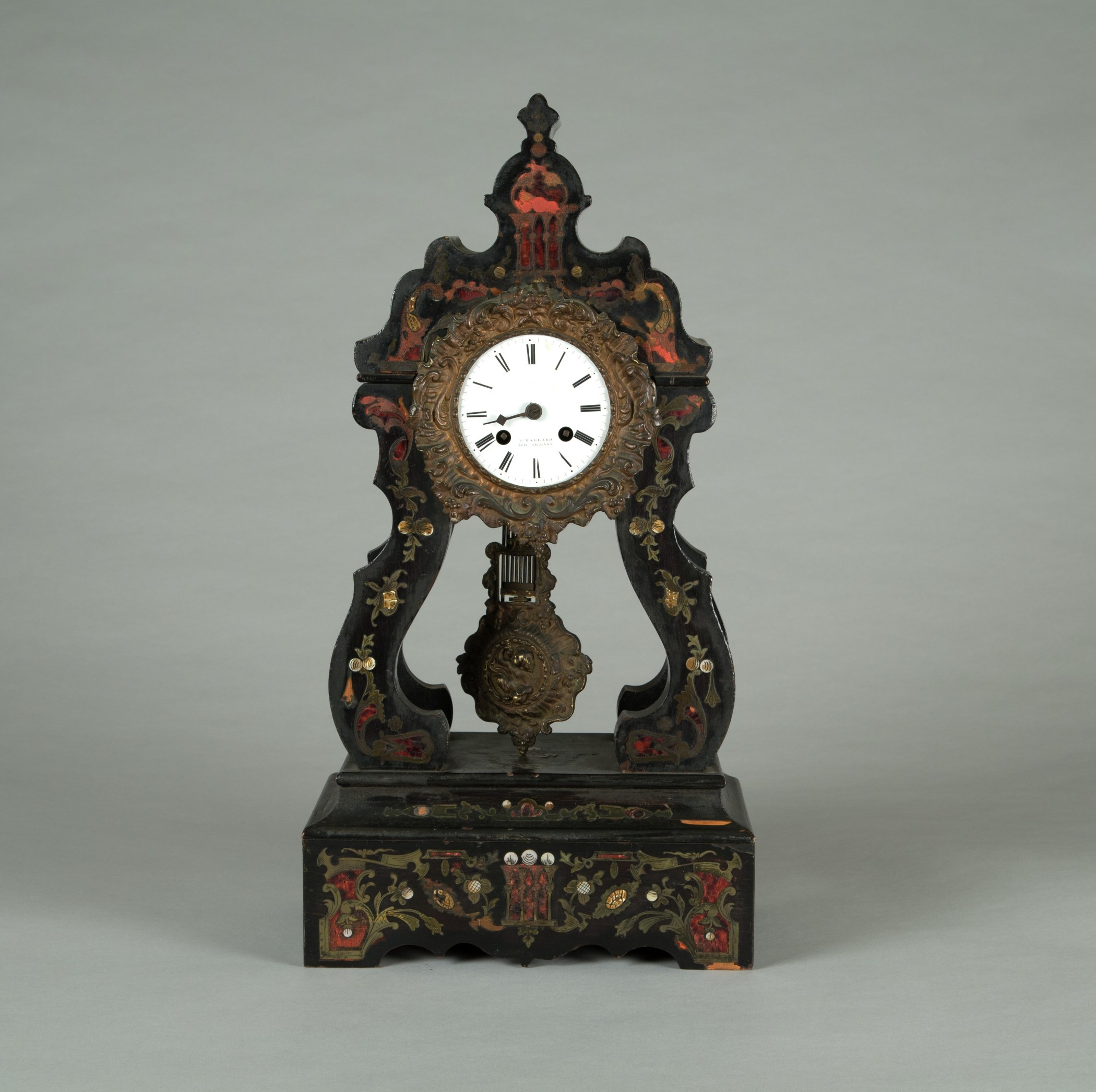 An ornate antique clock with detailed floral designs on a dark wooden frame, featuring a white clock face with Roman numerals and a decorative pendulum, set against a plain gray background.