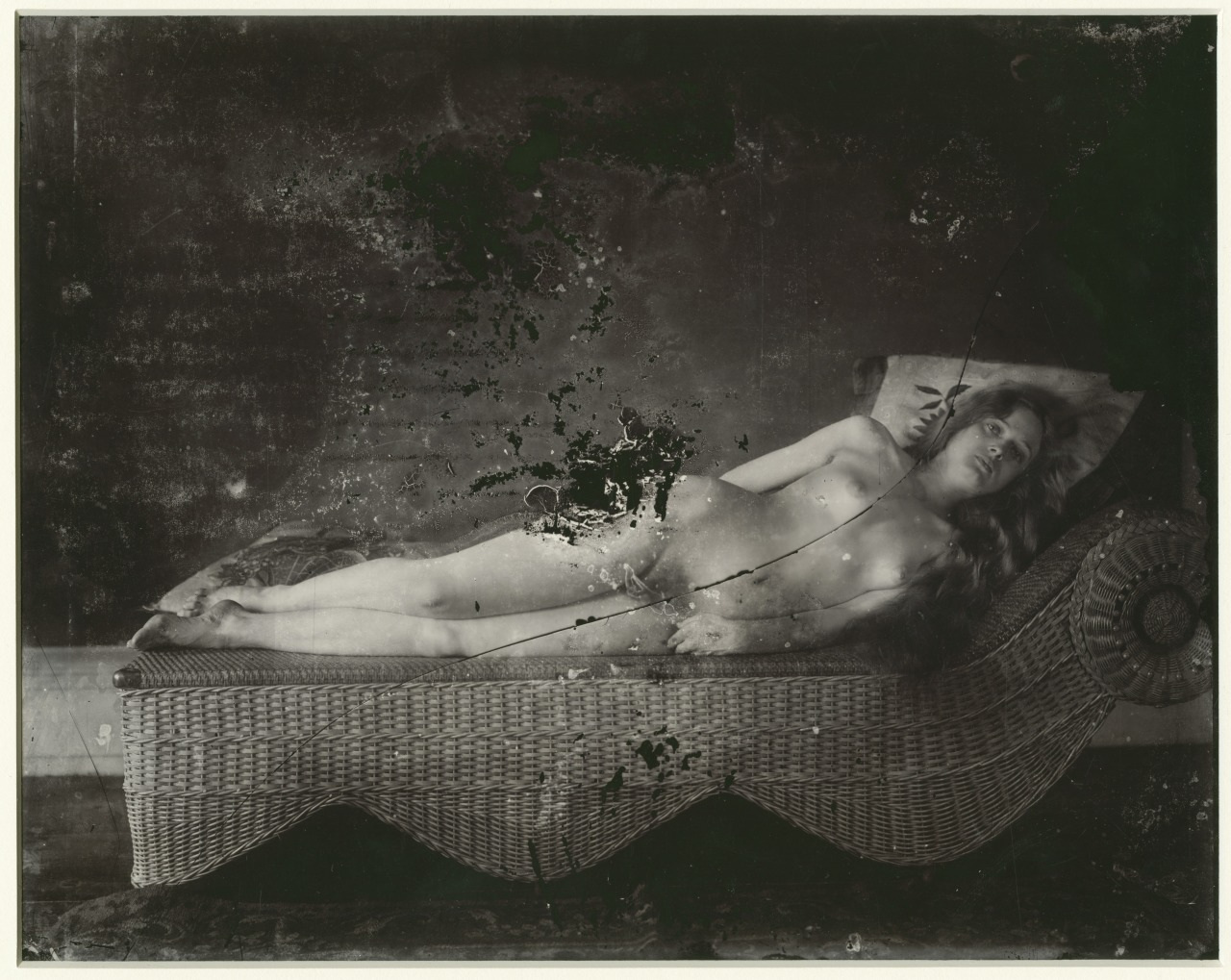 A black and white photograph of a person reclining on a woven chaise lounge, partially covered with a cushion. The image has a weathered and scratched appearance, adding a vintage feel to the scene.