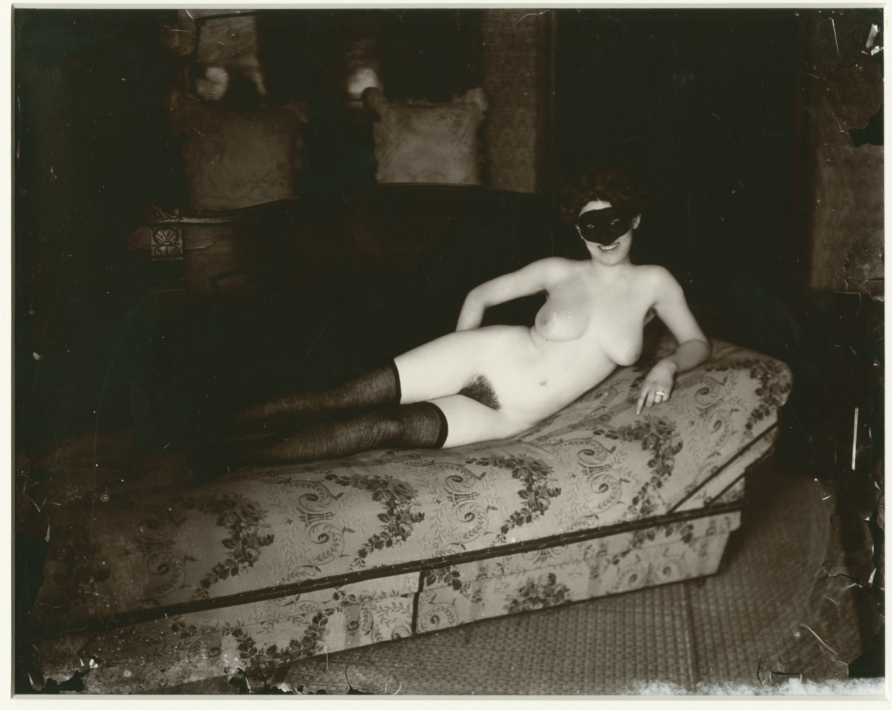 A sepia-toned vintage photograph of a person reclining on a patterned chaise lounge. The individual is wearing a black mask and dark stockings, with one arm resting on the back of the chaise. The room has ornate furnishings.