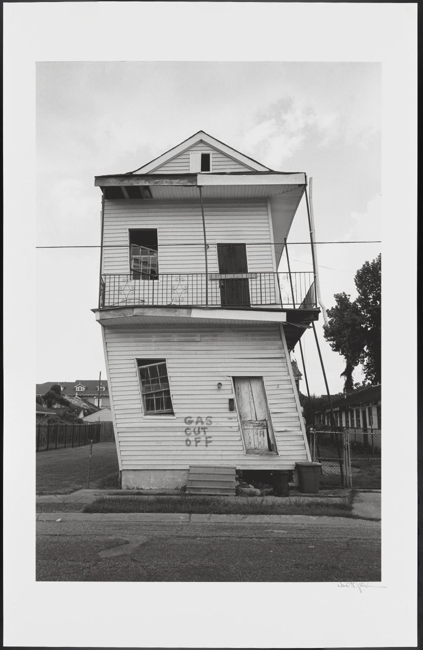 A leaning, two-story wooden house with visible damage and Gas Cut Off painted on the front. The structure appears abandoned, with broken windows and an unkempt yard, under a cloudy sky.