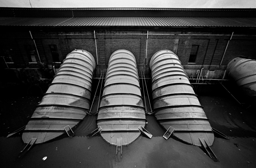 Black and white image of large, cylindrical structures submerged in water, leaning against a brick industrial building. The building has small windows along the top, and the scene appears still and industrial.