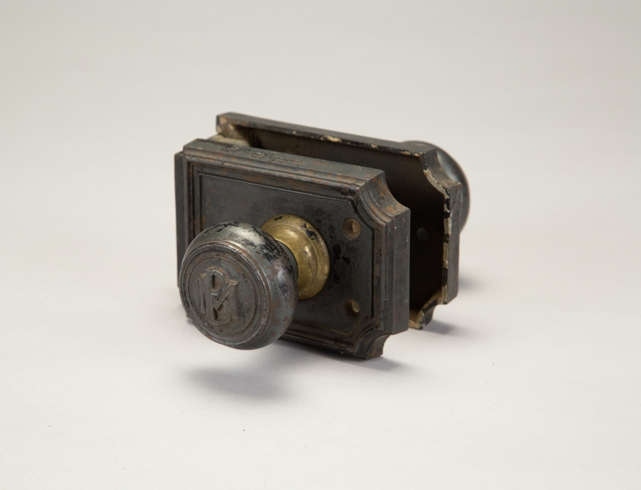 An antique metal doorknob and lock mechanism with decorative details, resting on a neutral surface. The knob features engraved initials or a monogram. The patina suggests age and use.