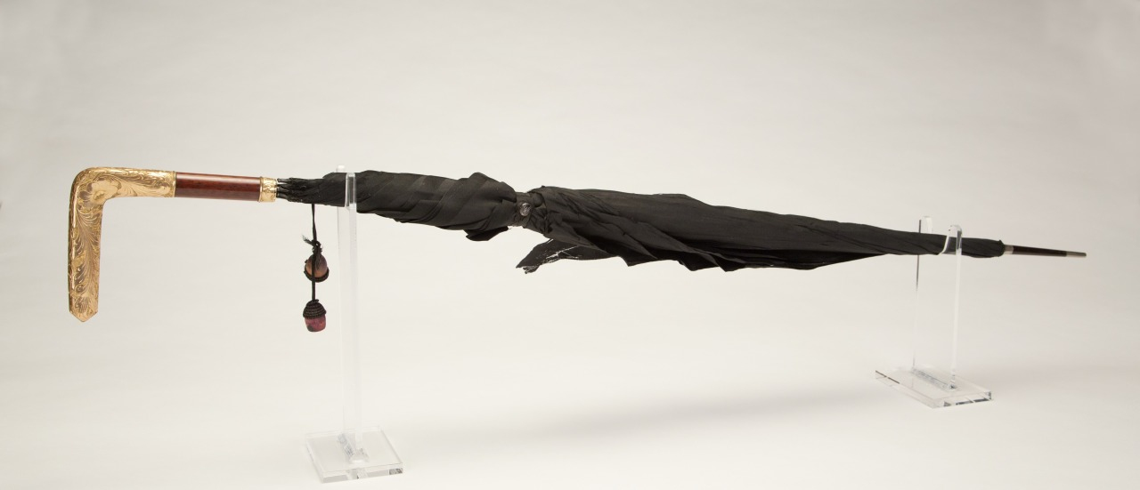 A closed black umbrella with a decorative gold handle, displayed horizontally on clear stands against a plain background.