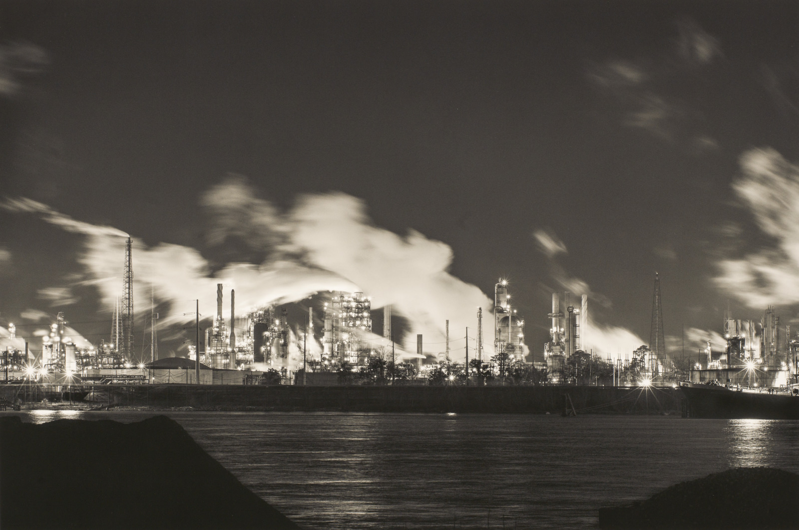 Nighttime industrial scene featuring a large factory complex with numerous chimneys emitting smoke or steam, set against a dark sky. The foreground includes a body of water, reflecting the factory lights.