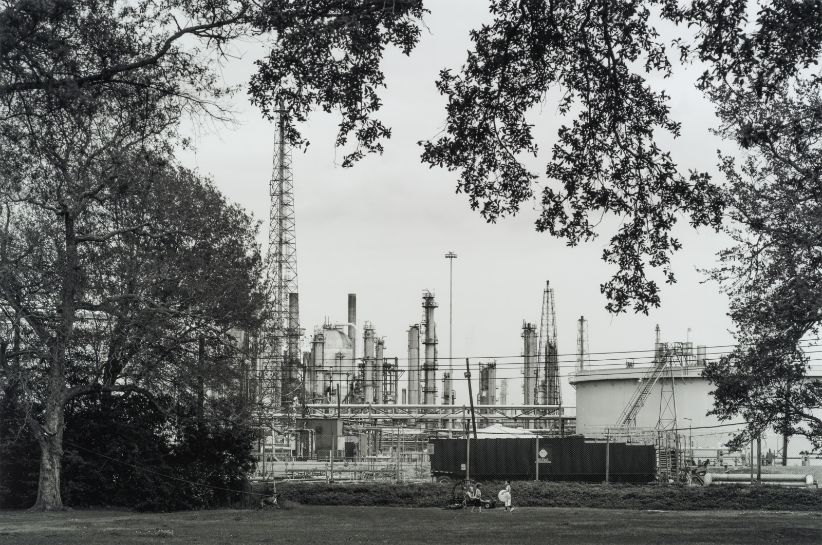 Black and white image of an industrial refinery complex with tall chimneys and storage tanks, seen through silhouetted tree branches. Two people sit on the grass in the foreground, providing contrast to the industrial backdrop.