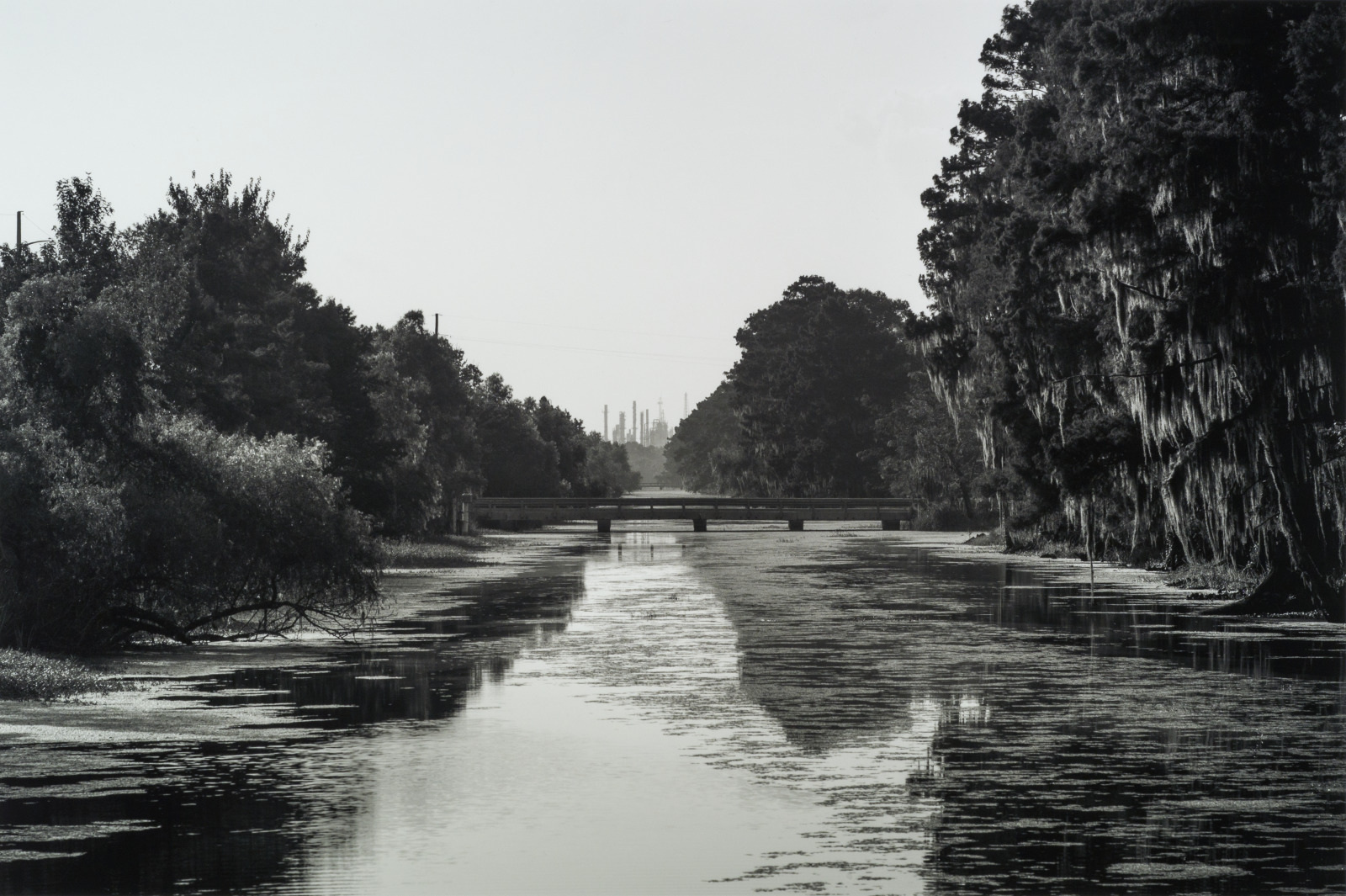 Black and white photo of a calm river flanked by trees, with Spanish moss hanging from branches. In the distance, a small bridge crosses over the water, and industrial buildings appear on the horizon, partially obscured by haze.