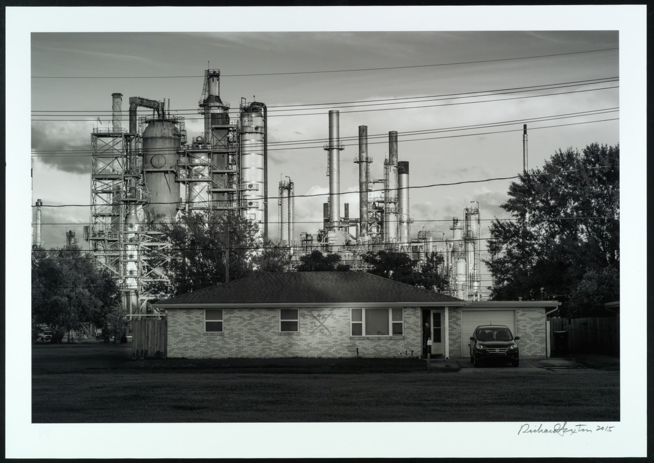 A black and white photograph of a small house with a car parked beside it. Behind the house, there is a large industrial refinery with tall towers and pipes, set against a cloudy sky. Power lines are visible in the foreground.