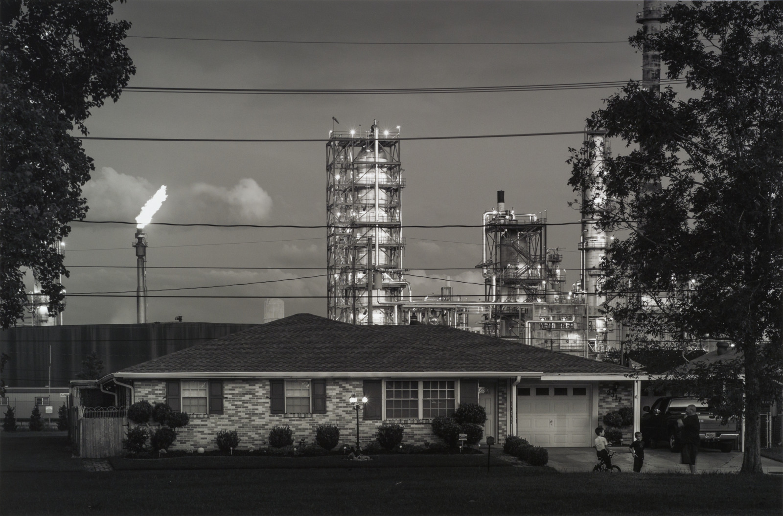 A small house stands in the foreground with an industrial plant behind it. Smoke and flames are visible from a stack. Trees frame the scene on either side, and power lines run across the image.