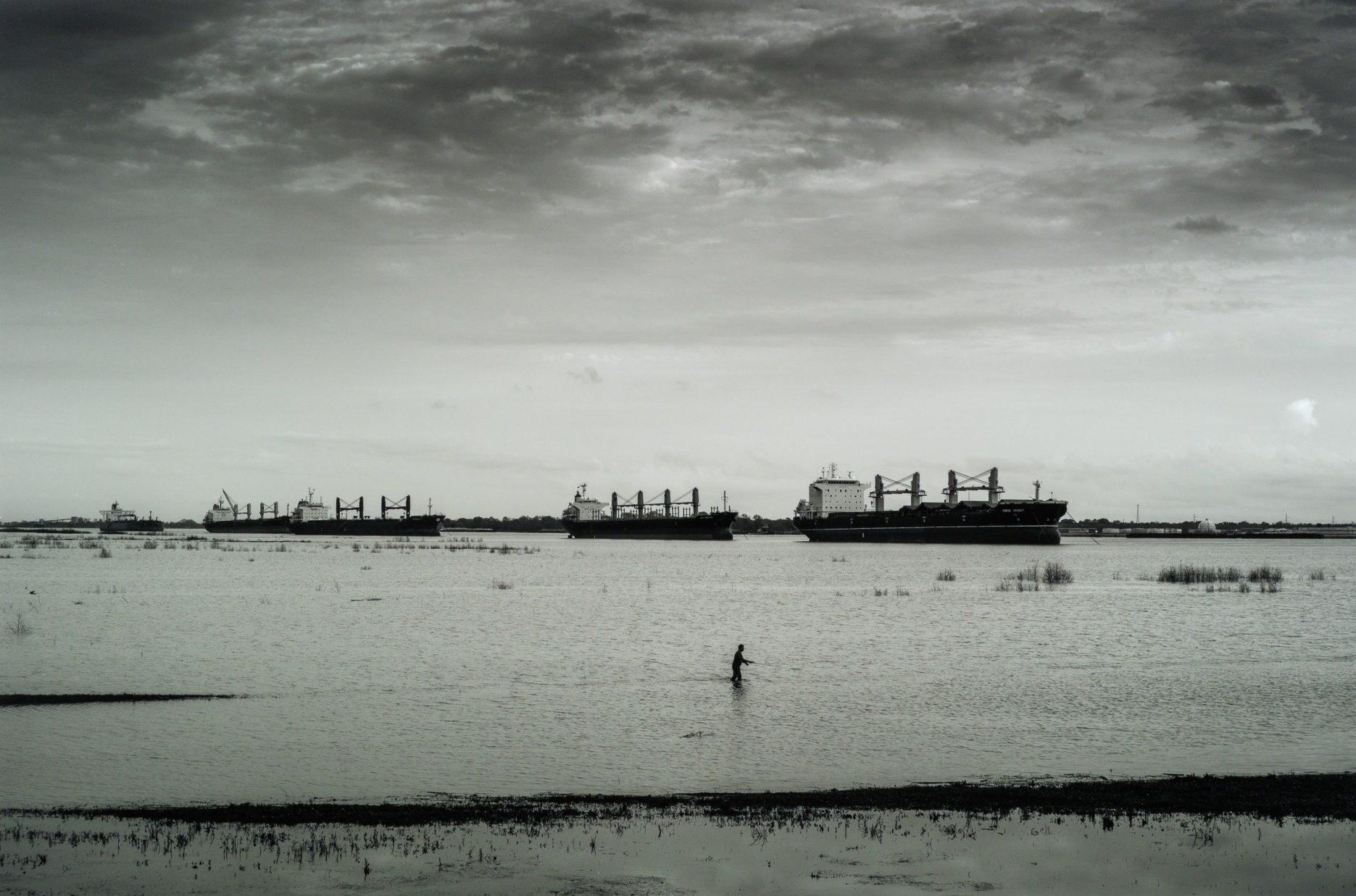 A black and white image shows a person wading in shallow water in the foreground. In the background, several large cargo ships are anchored under a cloudy sky, creating a stark contrast between the serene water and industrial ships.