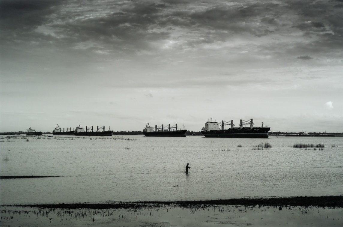 A serene, monochrome seascape with large cargo ships in the distance. A lone figure walks through shallow water in the foreground, under a cloudy sky, creating a peaceful yet dramatic scene.