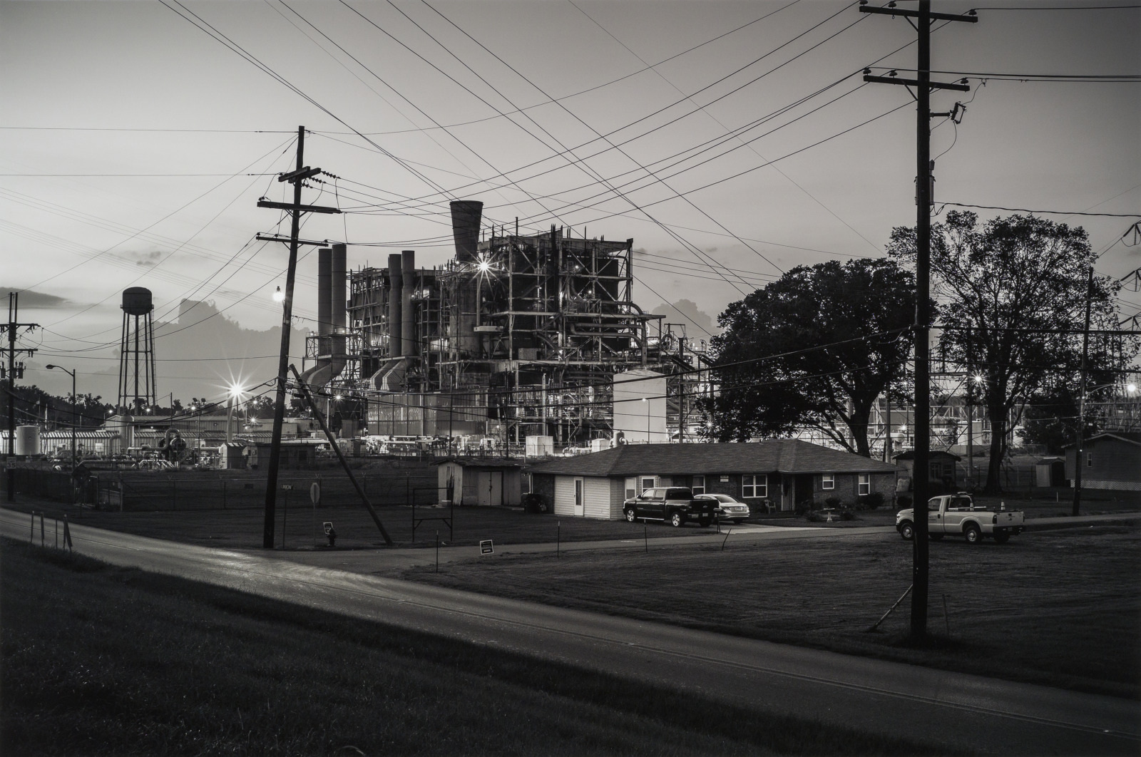Black and white photo of an industrial building with scaffolding, surrounded by power lines and a water tower. A small house and vehicles are in the foreground, with grass and a road leading toward the structure.