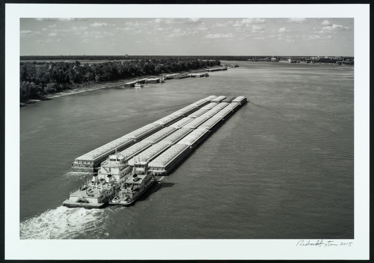 A long barge travels down a wide river, carrying multiple rectangular containers. The river is bordered by dense trees, and the sky is filled with scattered clouds. The black and white photo captures a sense of motion and tranquility.