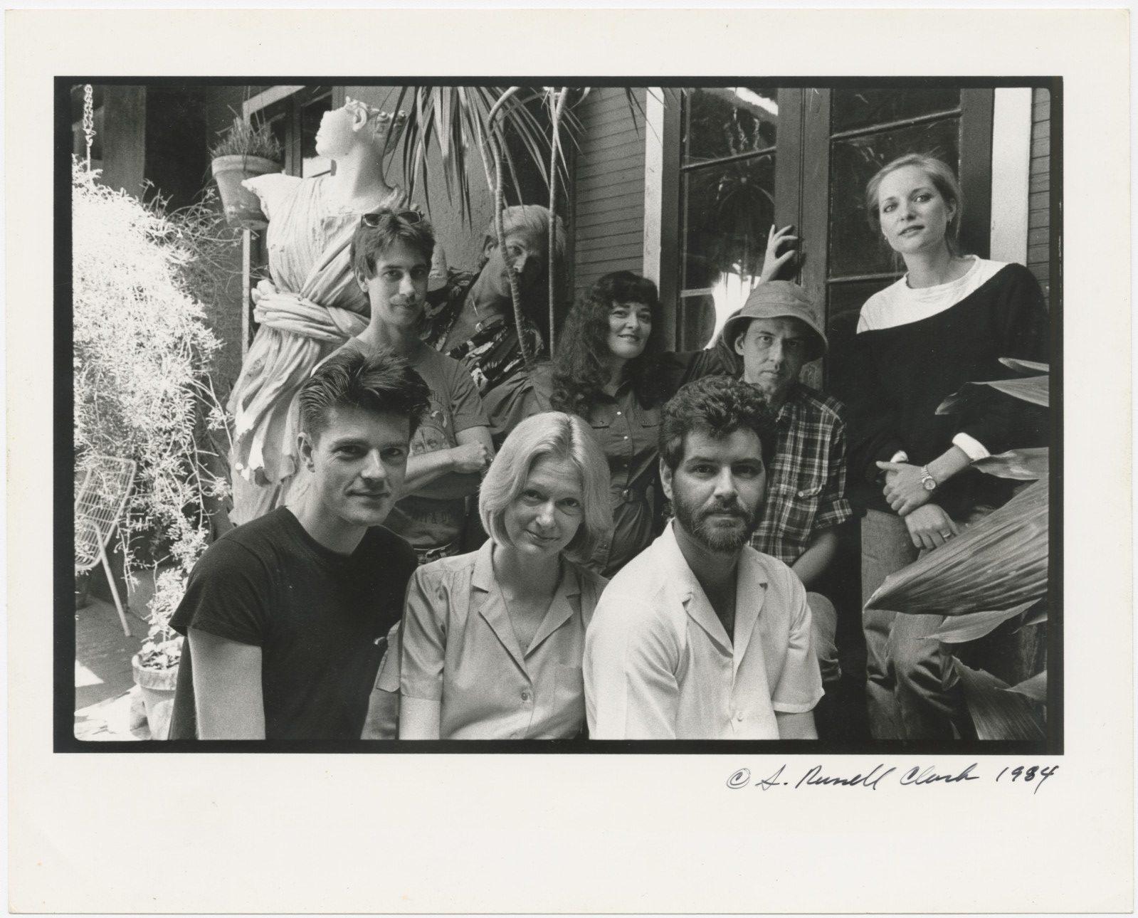 A black and white photo from 1984 showing a group of eight people posing together in a garden setting. They are casually dressed and some are smiling. Several plants and a statue are visible in the background.