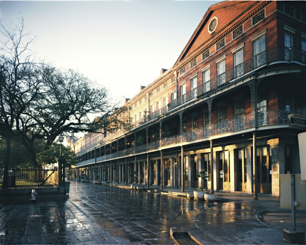 A street scene featuring a historic red brick building with intricate wrought iron balconies. The wet pavement reflects the warm sunlight, and leafless trees line the sidewalk.