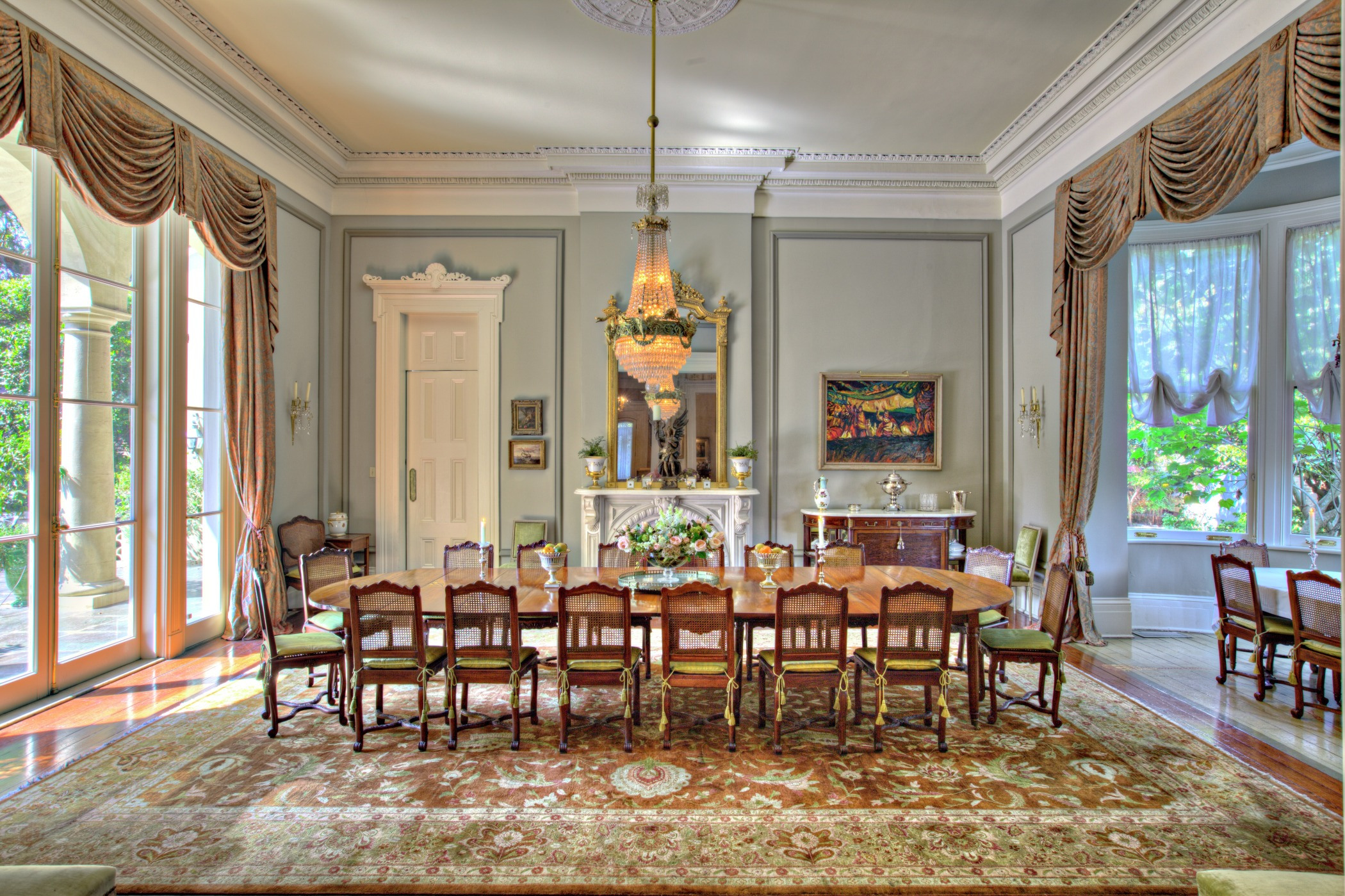 Elegant dining room with a large wooden table surrounded by chairs. A chandelier hangs from the ceiling, and a fireplace with a mirror is on one wall. The room features large windows, ornate curtains, and a decorative rug on the hardwood floor.