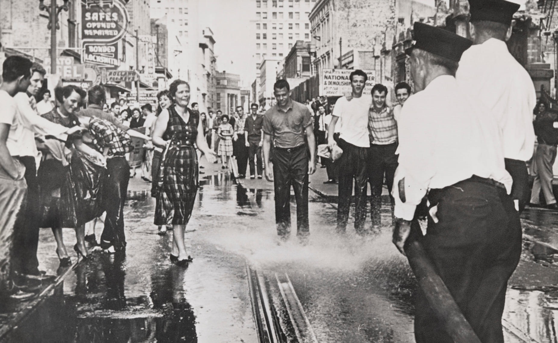 A black-and-white photo of a lively street scene with smiling men and women. People are walking through water sprayed by firefighters, while a crowd watches. The backdrop includes storefronts and city buildings, capturing a moment of joyful interaction.