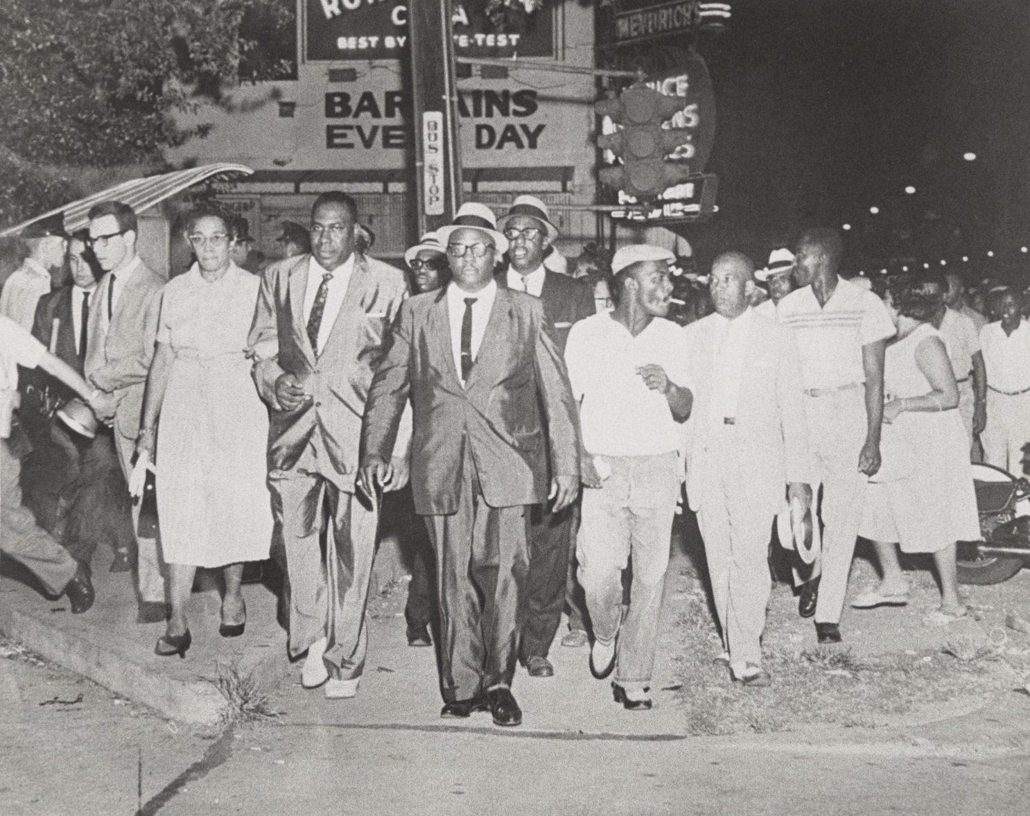 A group of civil rights activists, including men in suits and ties and women in dresses, march together on a street, surrounded by supporters. A sign for a restaurant is visible in the background. The scene is from a historical protest.