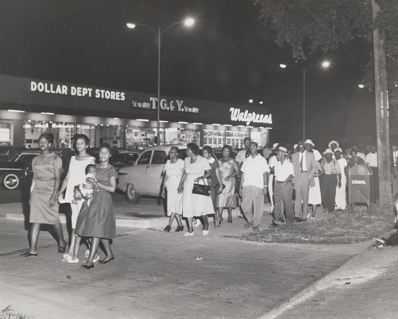 Civil rights protesters march past a shopping center. A Dollar Store and Walgreens can be seen in the background.
