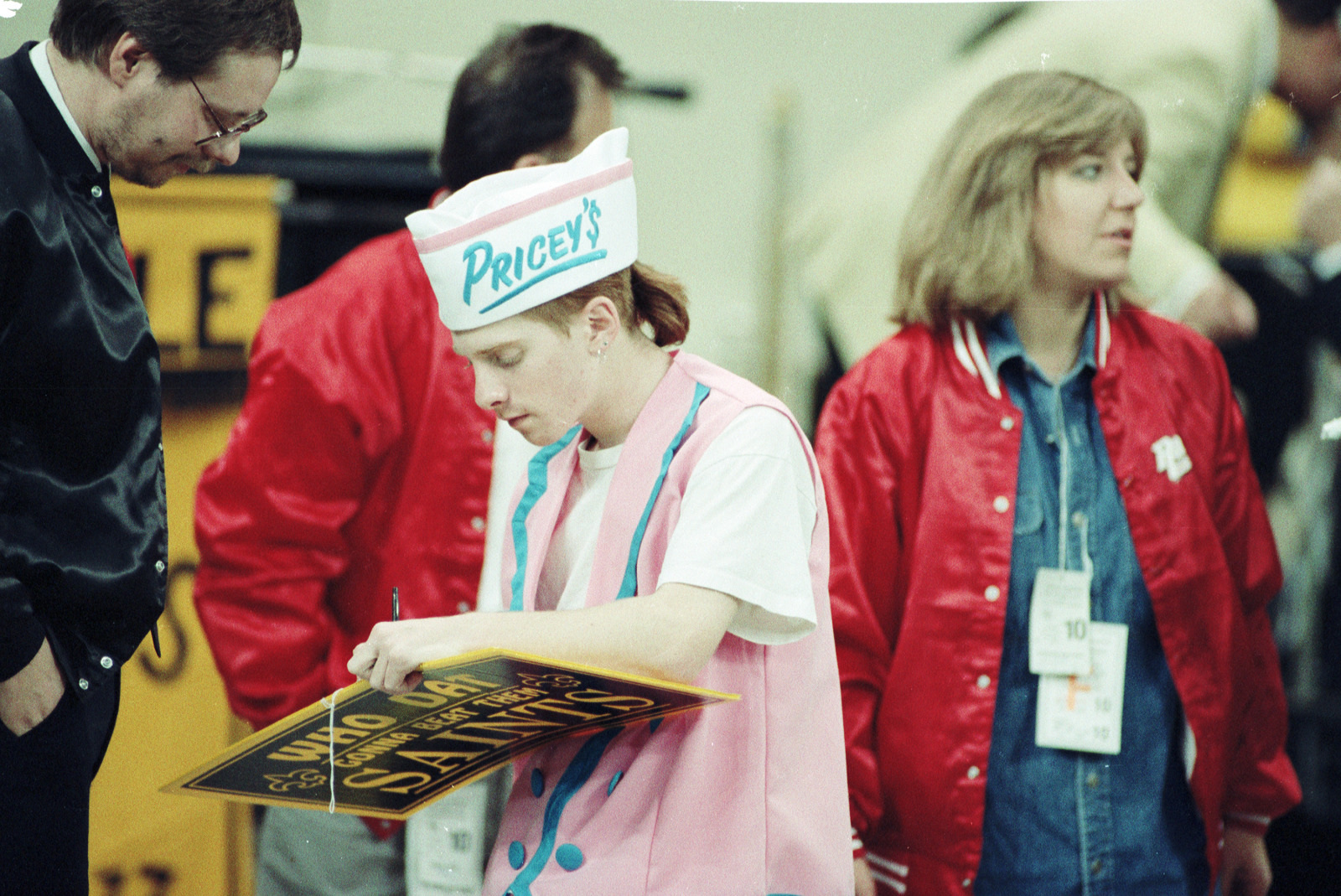 A person wearing a Priceys cap and pink uniform signs a black sign. They are surrounded by others in casual and sportswear. One person wears a red jacket. The scene suggests a busy event or gathering.