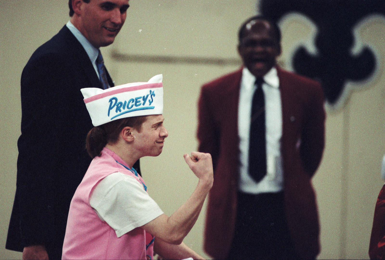 A person wearing a Priceys paper hat and pink uniform celebrates with a fist pump indoors. Two men in suits are in the background, one laughing.