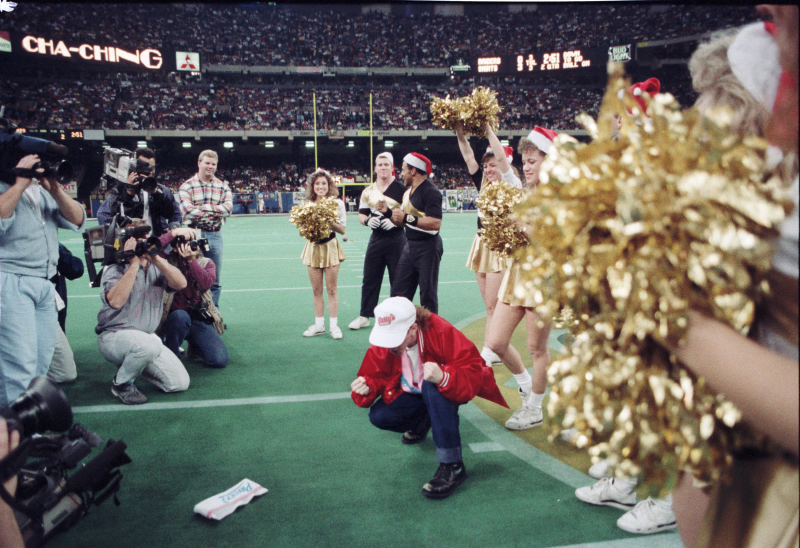 A person in a red jacket and white pants kneels on a football field, surrounded by cheerleaders with gold pom-poms and photographers capturing the scene. The cheerleaders wear red Santa hats. The stadium is filled with spectators.